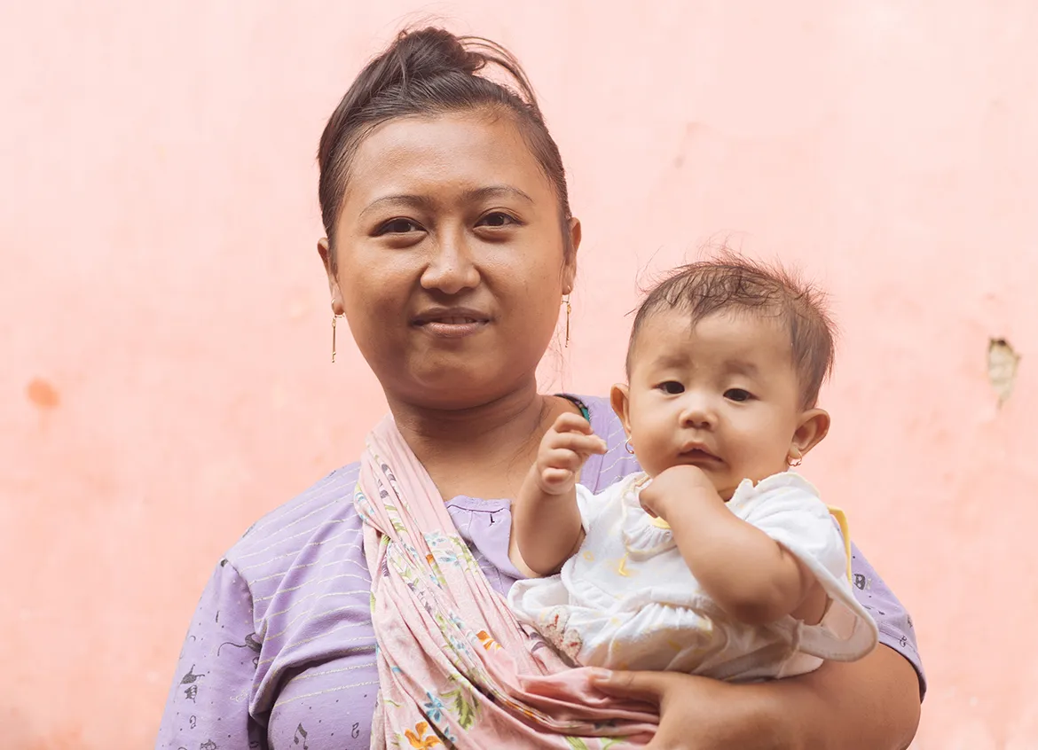 A woman smiling while holding her young child in front of a pink wall in Indonesia.