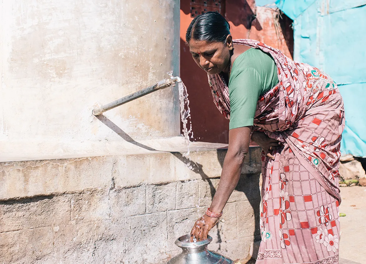 Woman filling silver water vessel from spigot outside her home.