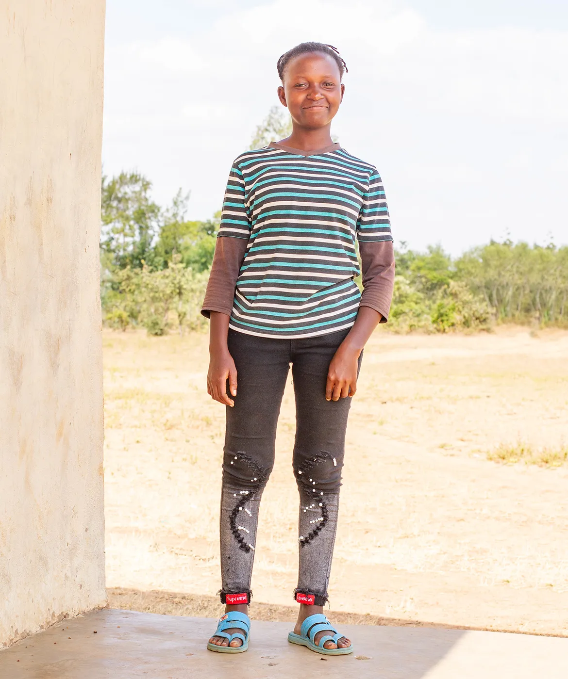 A young woman named Ann standing near a tan building, smiling while wearing a striped shirt and blue sandals.