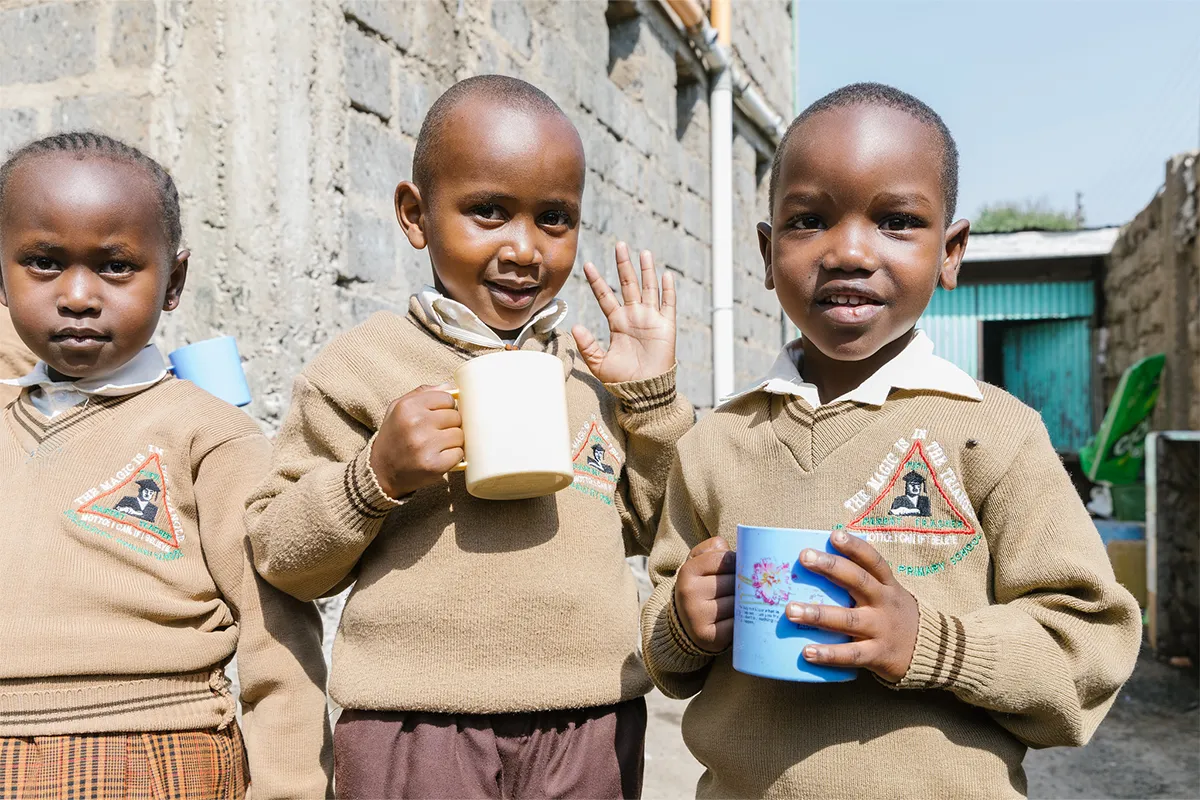 Three young children in school uniforms wave holding cups of safe water.