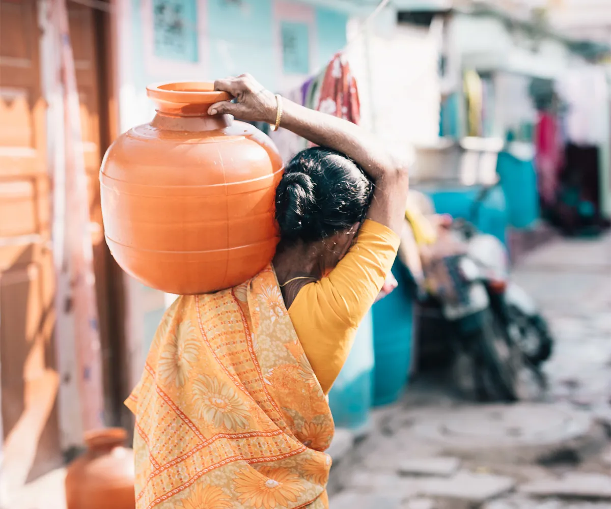 Woman walking down the street carrying an orange water vessel on her left shoulder.