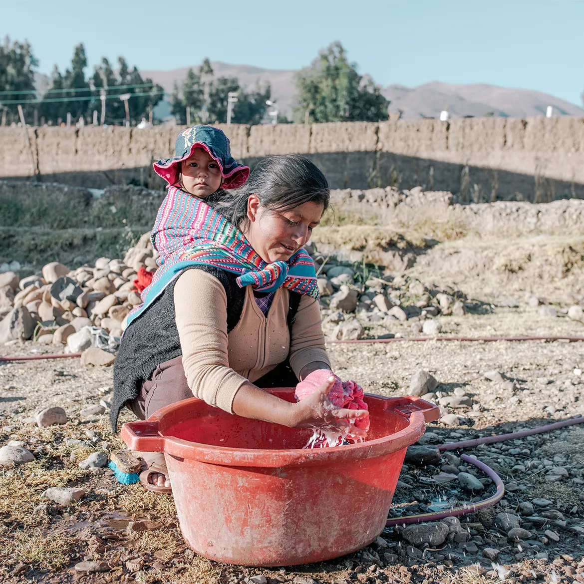 Woman in Peru doing laundry in a red bucket with her child tied around her back.