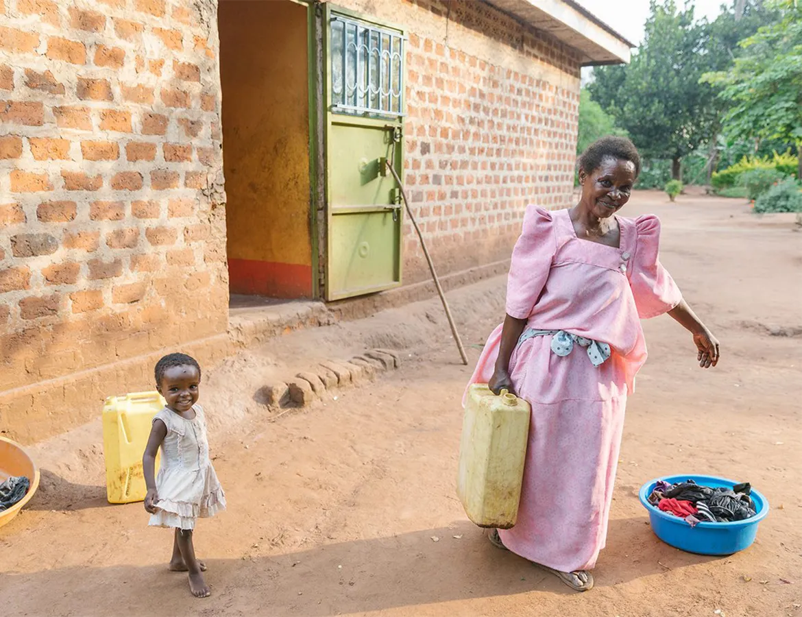 Woman holding a water vessel for laundry while standing next to a young girl outside of a brick building.